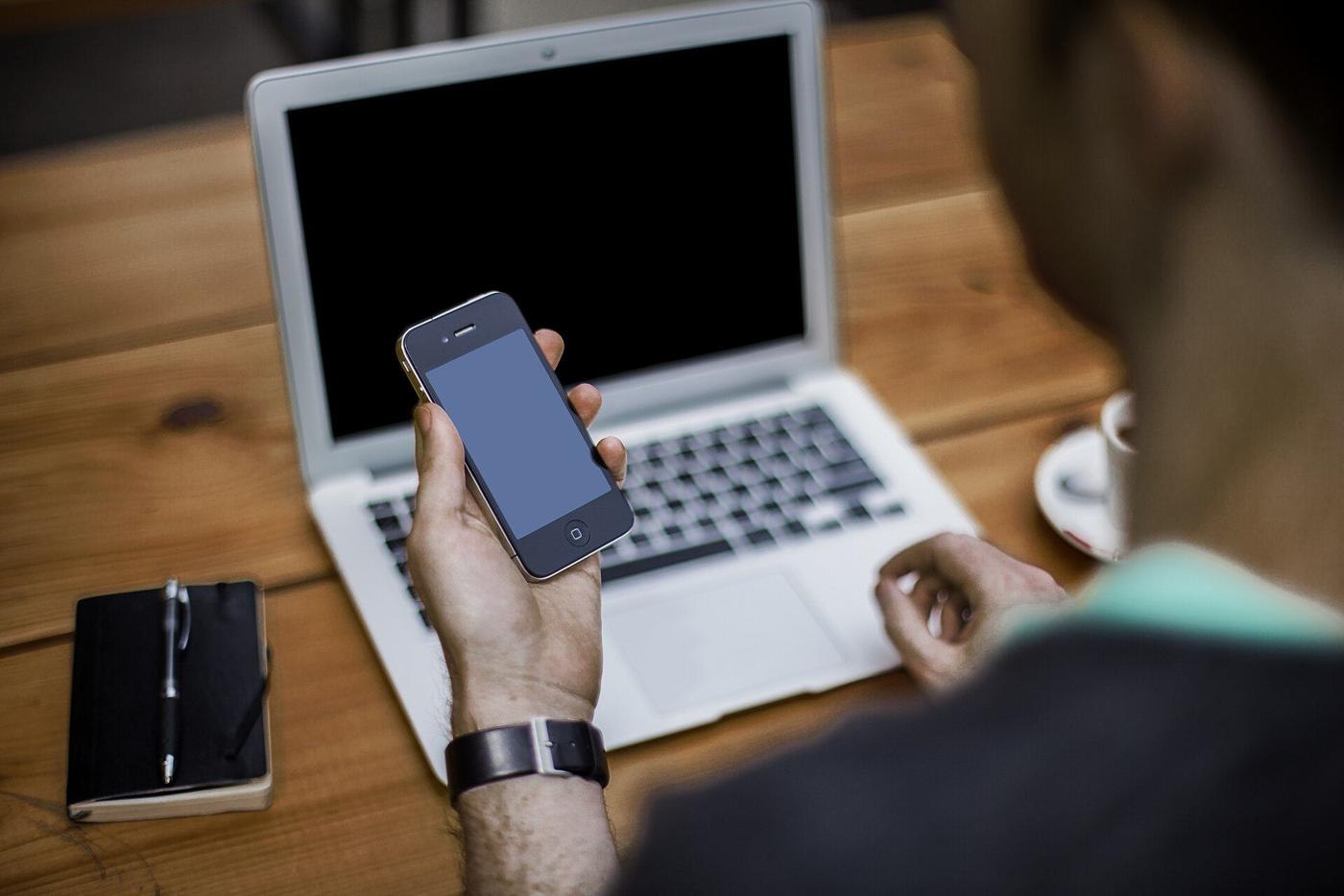 A person holding a phone in front of a laptop on a wooden desk
